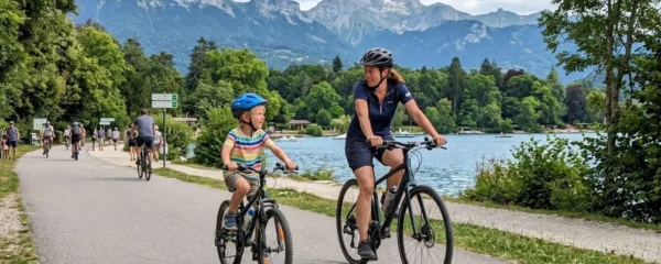 An adult and child cycling side by side on a smooth paved greenway with Lake Annecy's turquoise waters visible through trees in the background